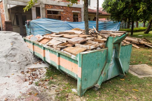 A large green industrial skip filled with a substantial pile of broken wooden planks, panels, and debris, situated on a grassy area adjacent to a construction or renovation site in a residential neighbourhood. The wood varies in size and thickness, with some pieces showing weathered textures and natural wood tones, while others have painted or treated surfaces. The skip is positioned next to a dirt pathway and is partially surrounded by scattered leaves and soil. Behind the skip, there is a partially built brick structure with some blue tarp covering part of its upper section, suggesting ongoing construction. To the right, a mature tree and a row of residential houses with gardens are visible, contributing to the suburban context. The overall scene is illuminated by daylight, with the environment indicating domestic or local building rubbish typical of a garden or property clearance in the Blackwall area, serviced by Garden Clearance Blackwall.