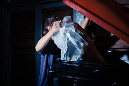 A person with short, dark hair and wearing a dark shirt is lifting a transparent plastic bag filled with various types of rubbish, including paper and possibly garden waste, into a large black wheelie bin located outdoors. The scene appears to be set in a driveway or yard, with the background consisting of dark wooden panels and metal framing. The person is positioned on the left side of the image, with their arms raised, and the bin is partially visible at the bottom right. The lighting suggests an overcast or shaded environment, typical of regular waste collection areas. This image is relevant to rubbish removal services, such as those provided by Garden Clearance Blackwall, which operates in the Blackwall area, supporting local waste management needs in east London.