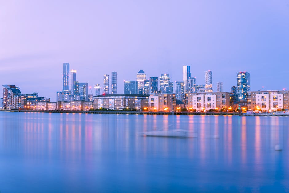 A cityscape featuring several modern high-rise buildings with glass facades reflecting the blue sky and clouds. In the foreground, there is a body of water with a dark metal railing running along its