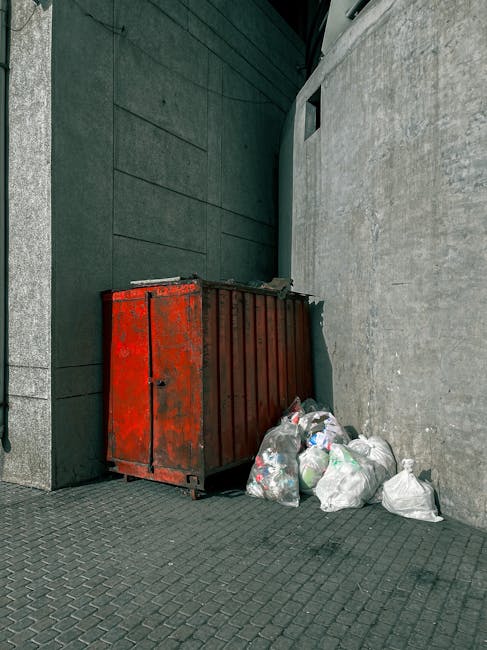 A red metal skip with visible rust and minor dents is positioned against a dark green wall adjacent to a light gray concrete wall in an urban outdoor setting. The skip's surface appears weathered, wit