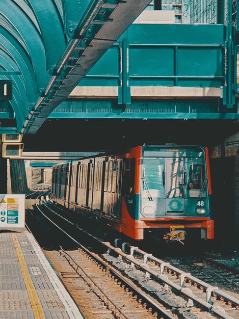 A modern urban scene featuring a red train crossing a bridge over a river in a city with tall, contemporary glass and steel skyscrapers. The buildings vary in height and architectural style, with some